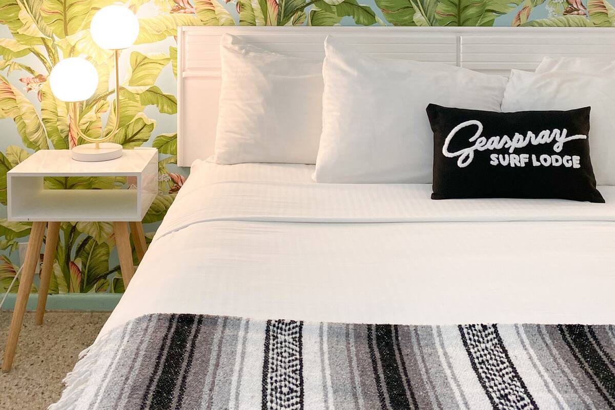 A bedroom with a bed, white bedding, a nightstand with a lamp, and a pillow with text.