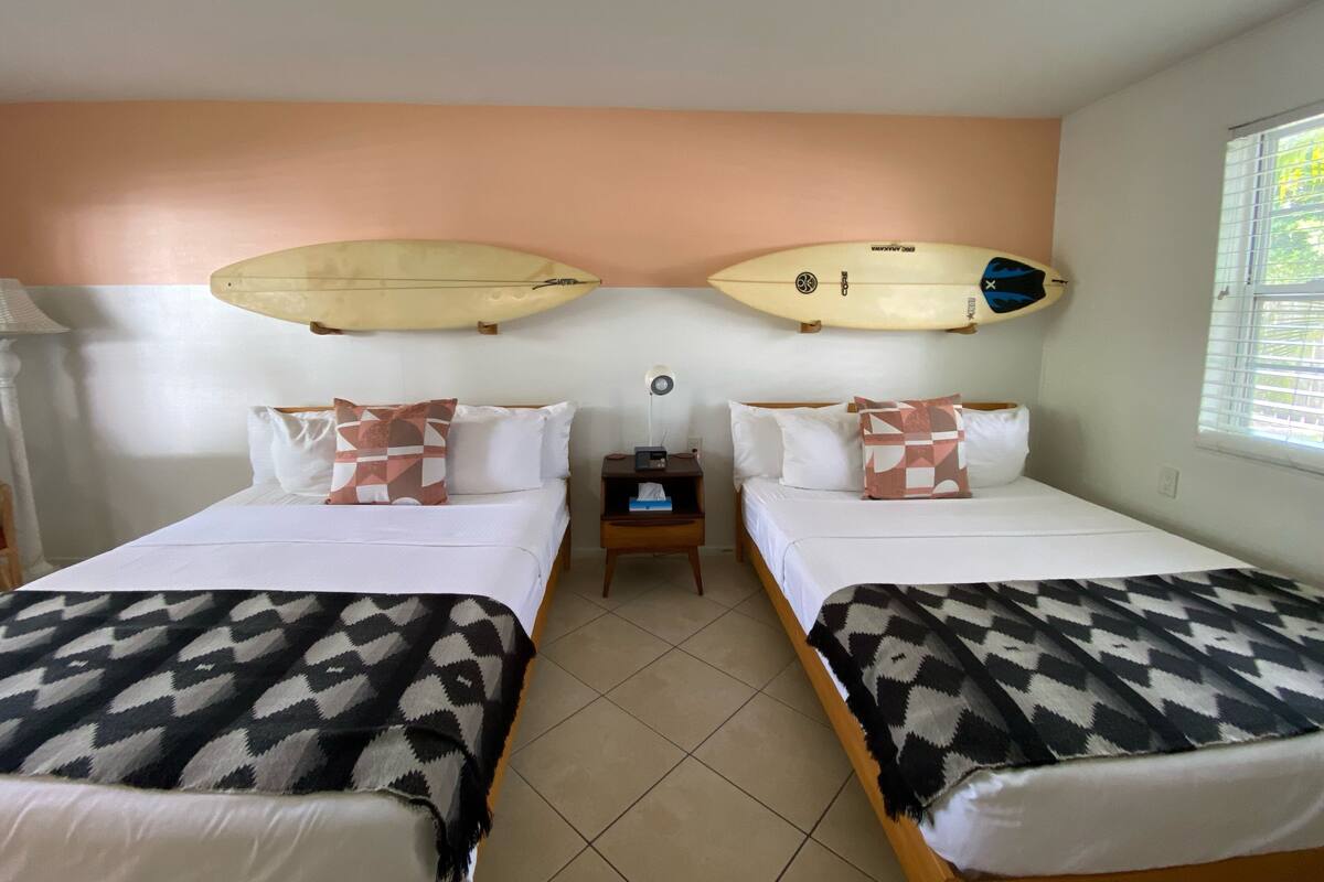 A hotel room with two beds, each with patterned blankets and pillows, a nightstand with a lamp, and surfboards mounted on the wall.