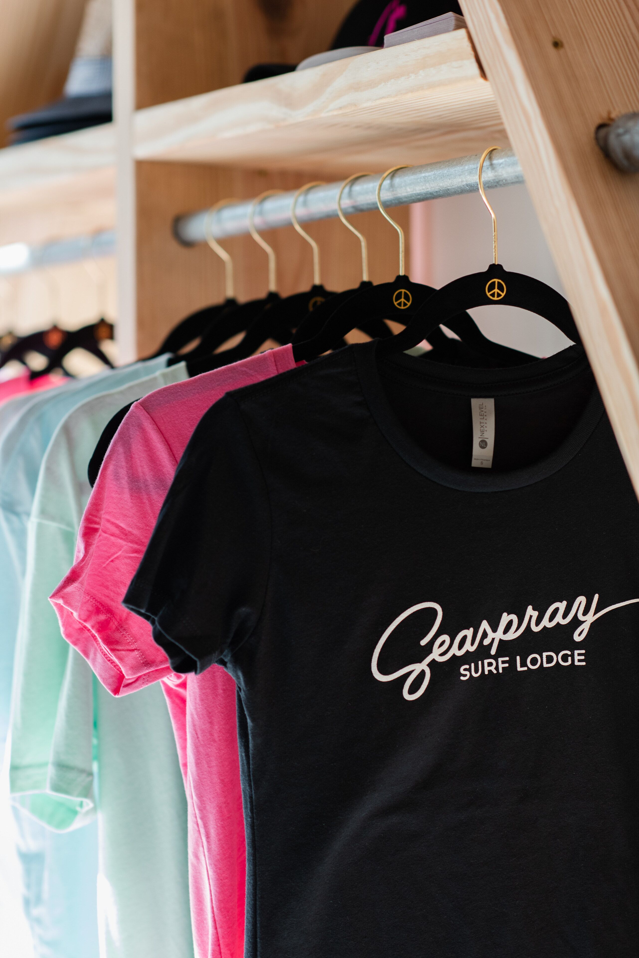 Una rastrelliera con magliette di diversi colori, tra cui una nera con la scritta 'Geaspray Surf Lodge'.