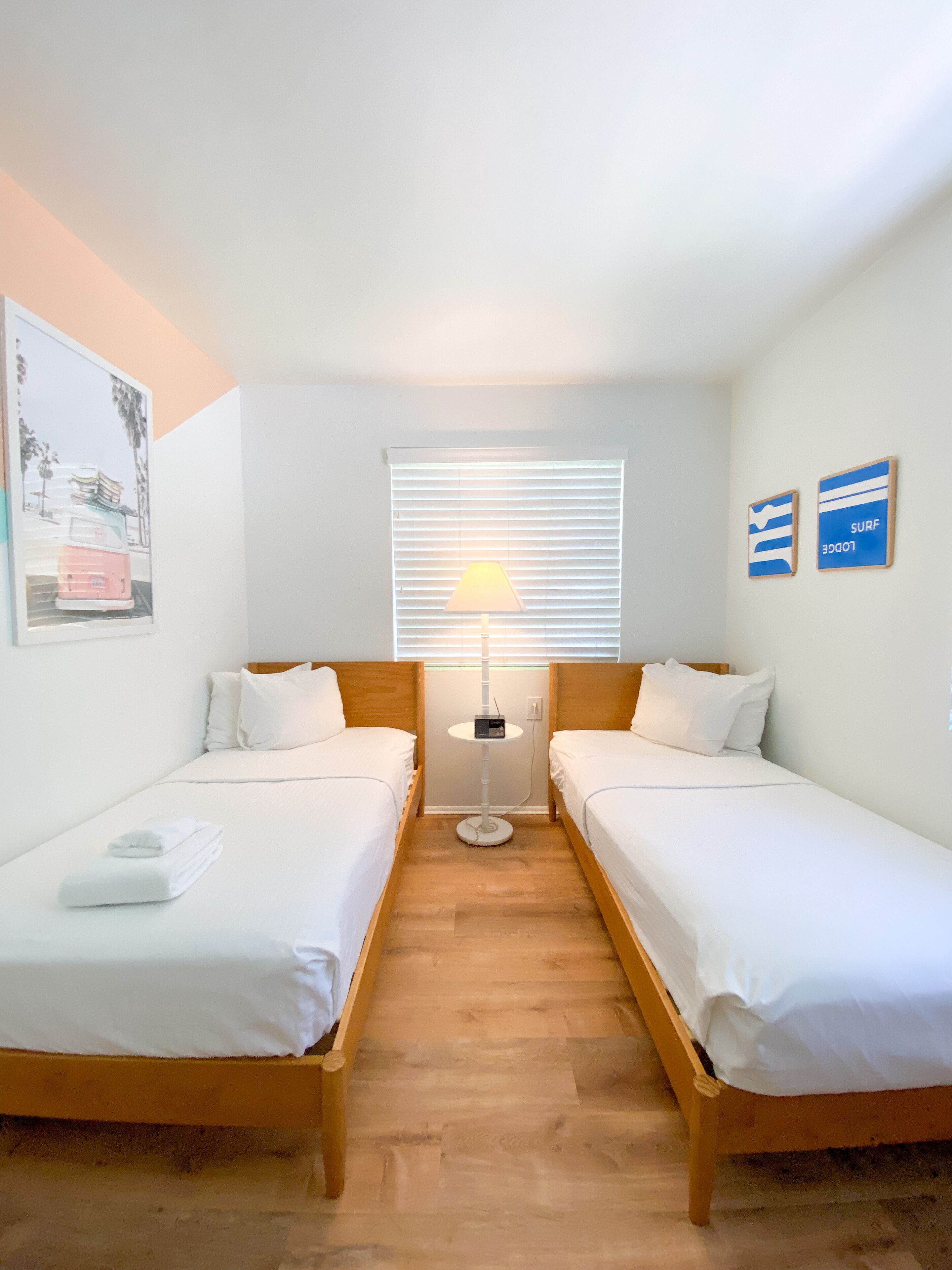 A room with two single beds, a standing lamp, a window with blinds, and two wall-mounted signs.