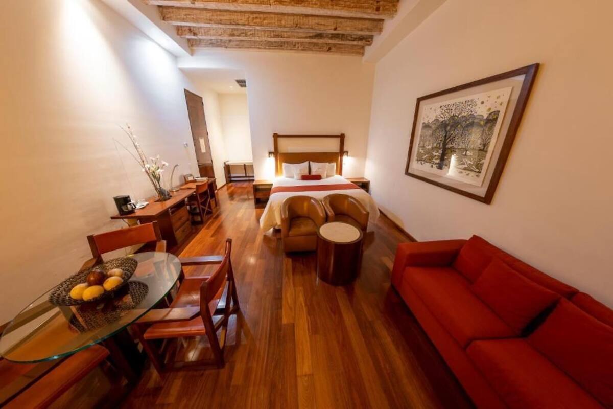 A cozy room with a wooden floor, a red sofa, a glass dining table with fruit, a bed with a wooden headboard, and a framed picture on the wall.