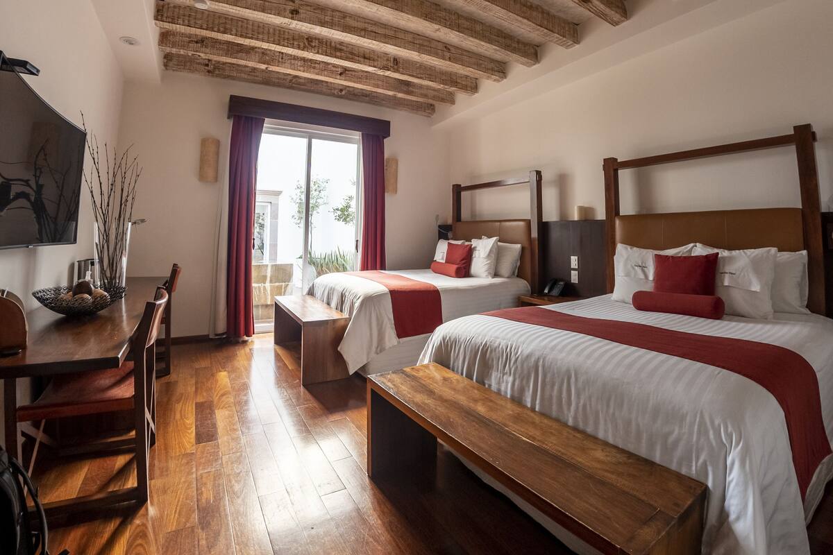 A hotel room with a wooden ceiling, two beds with red and white bedding, a wooden desk, and a bench.