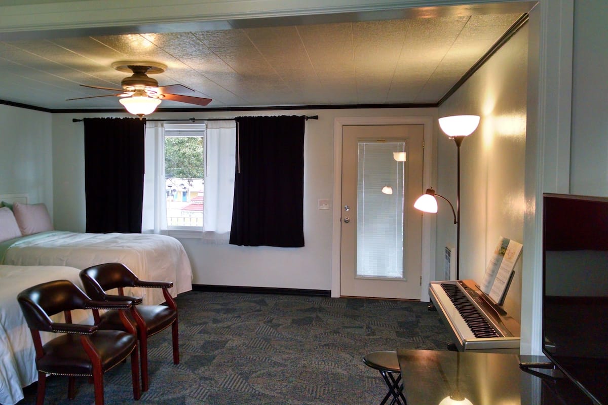 A hotel room with two beds, a ceiling fan, a piano, and a small table.