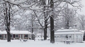 Un parc cobert de neu amb una glorieta, bancs i un edifici amb teulada nevada.