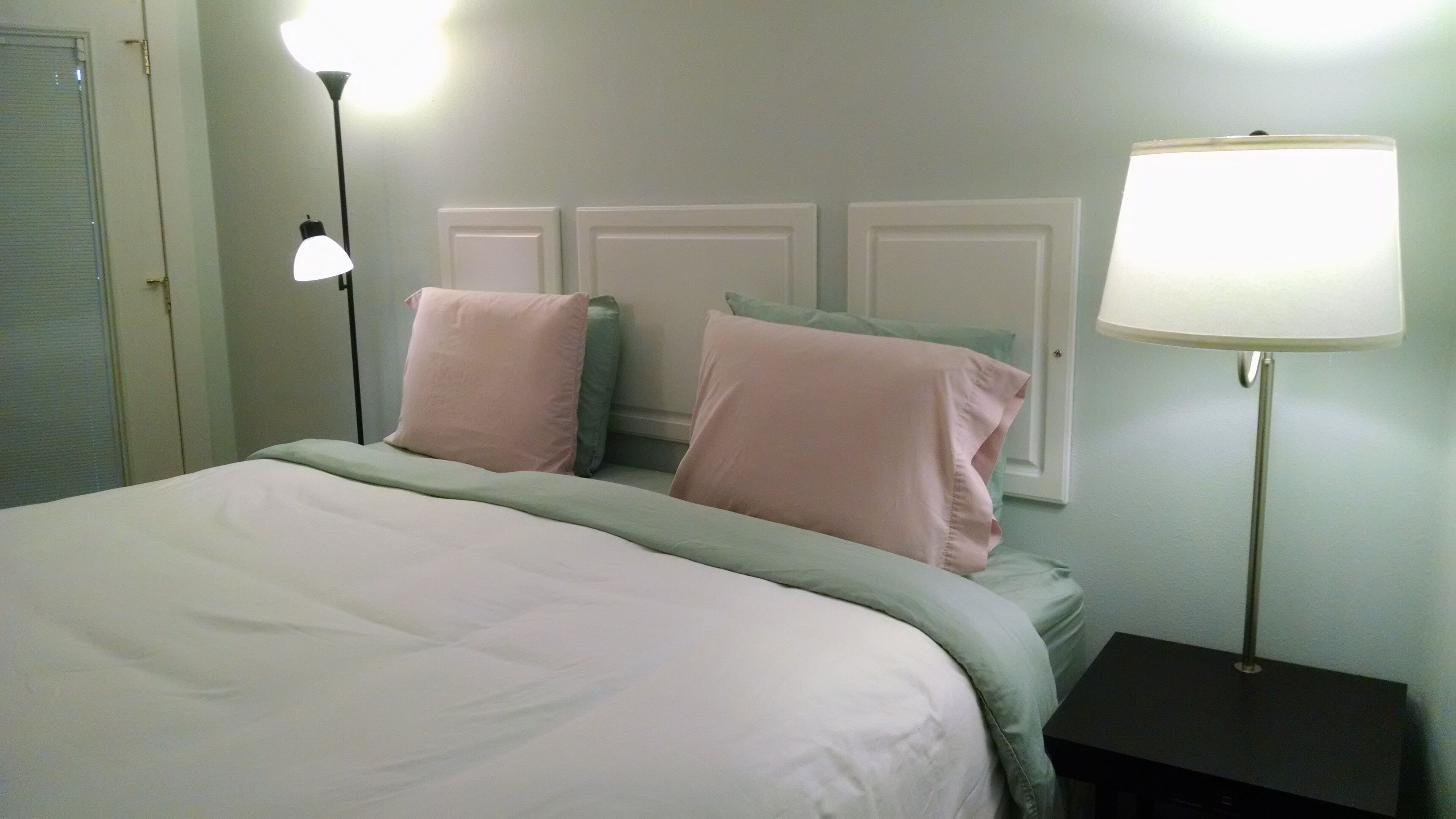 A neatly made bed with a white and green bedspread, two pink pillows, and a bedside lamp on a small table.