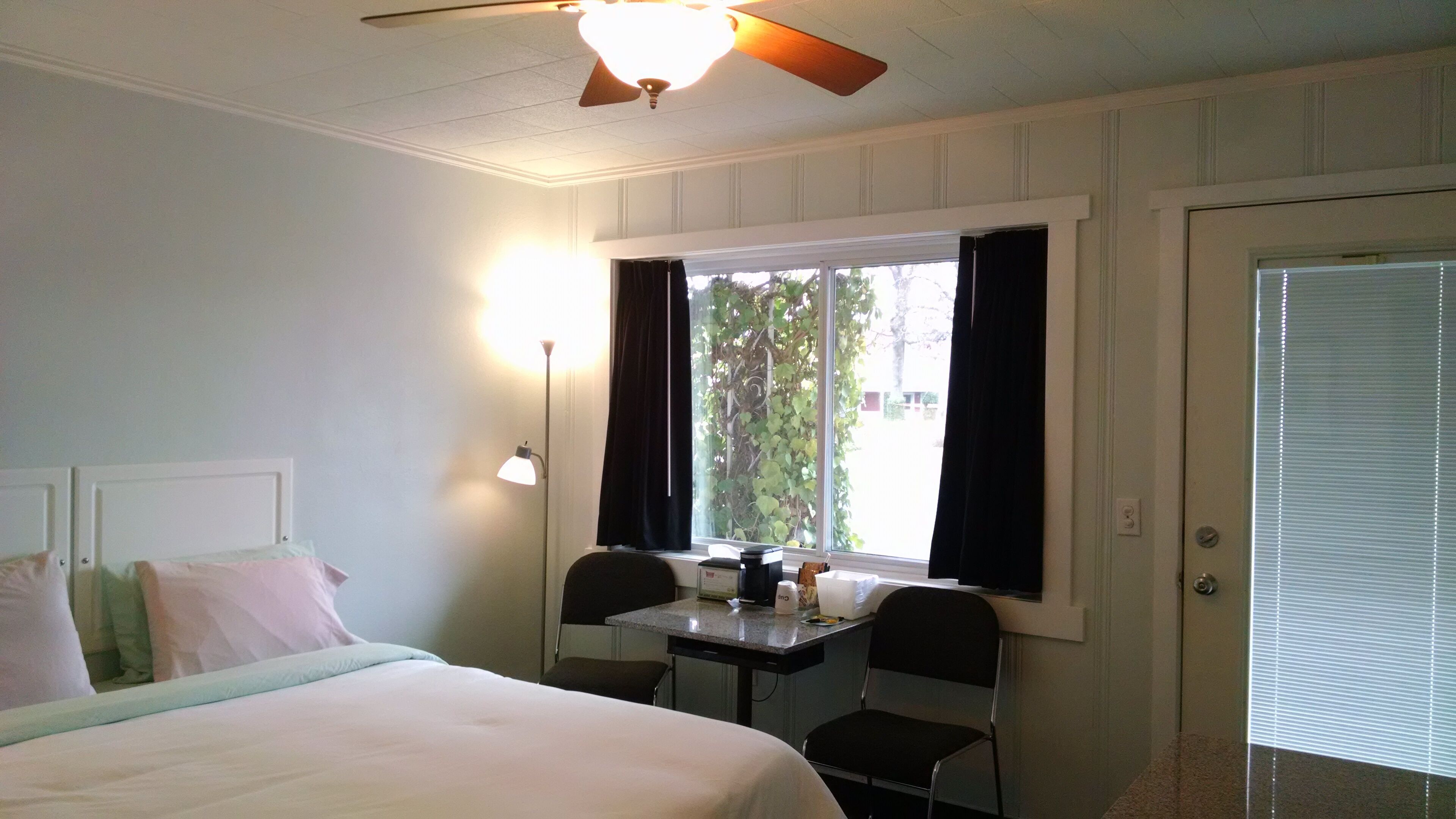 A hotel room with a bed, a desk with two chairs, a ceiling fan, a window with curtains, and a door.