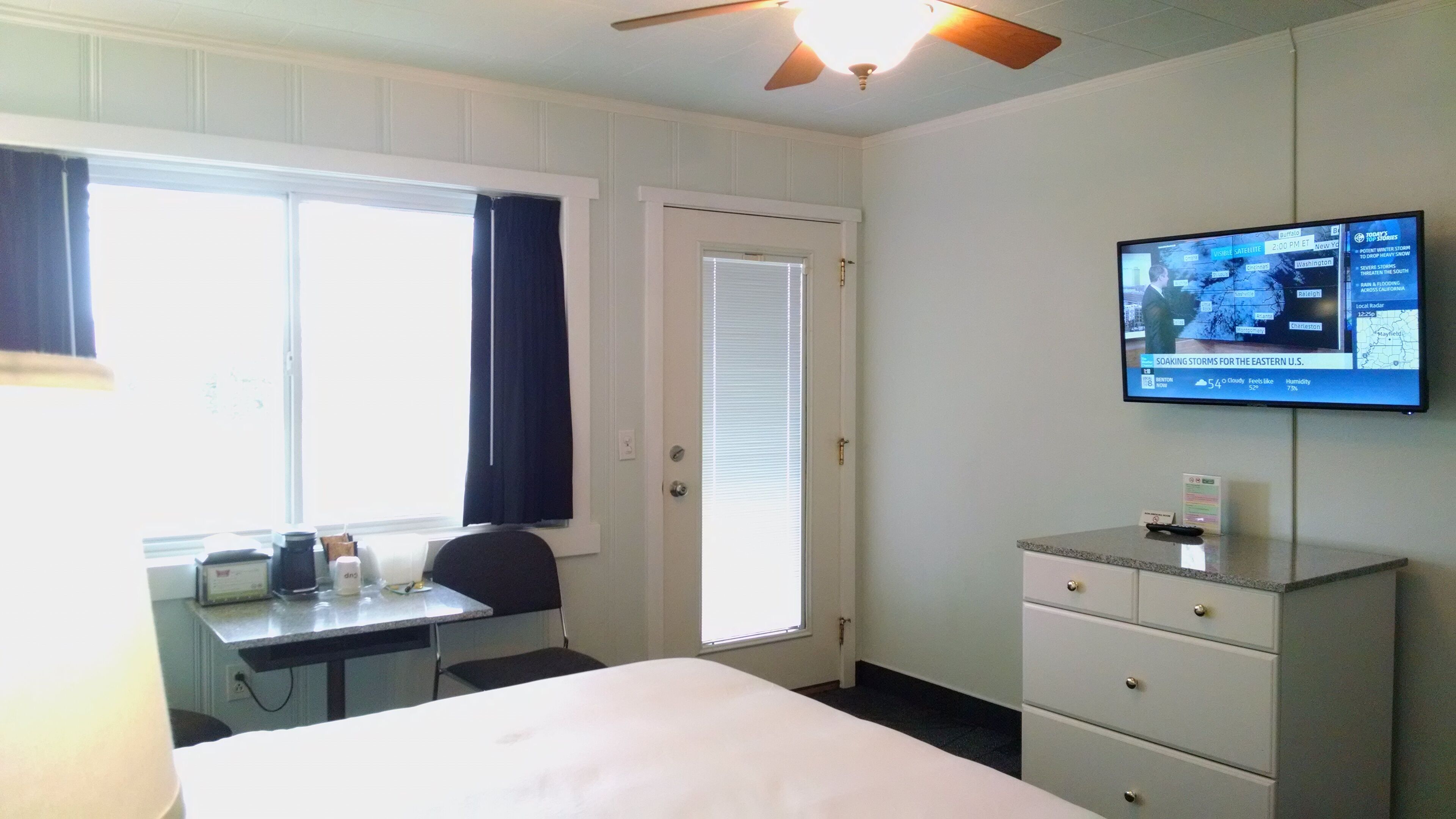 A hotel room with a bed, a desk, a chair, a television mounted on the wall, and a ceiling fan.