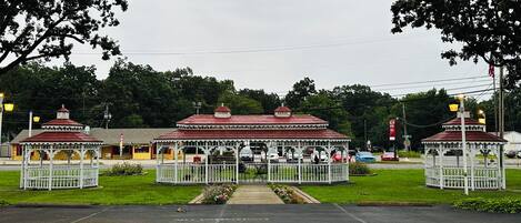 Une structure de type belvédère, avec un toit rouge et des bordures blanches, entourée d’une clôture blanche et d’un petit jardin.