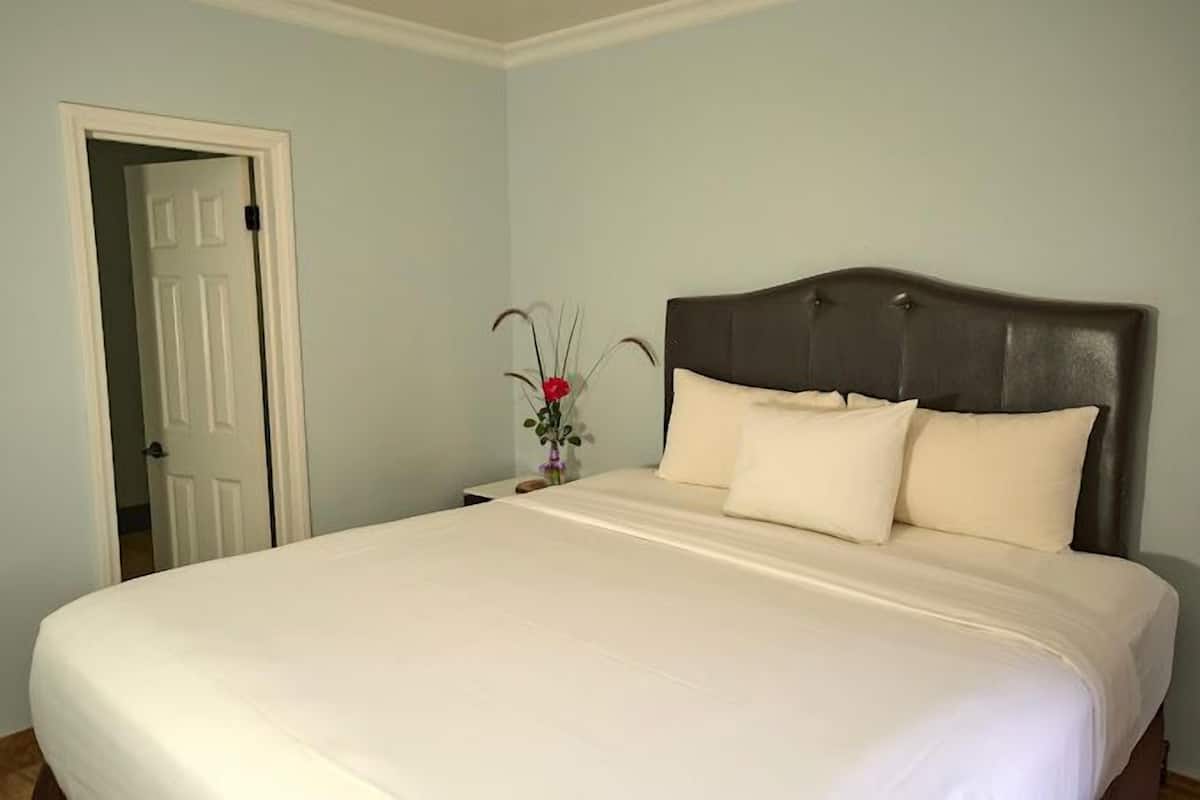 A neatly made bed with a dark headboard, a white door to the left, and a small table with a vase of flowers.