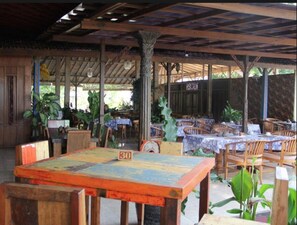 An outdoor restaurant with wooden tables and chairs, covered by a wooden roof, and surrounded by potted plants. - Gazebo Beach Hotel (Denpasar)