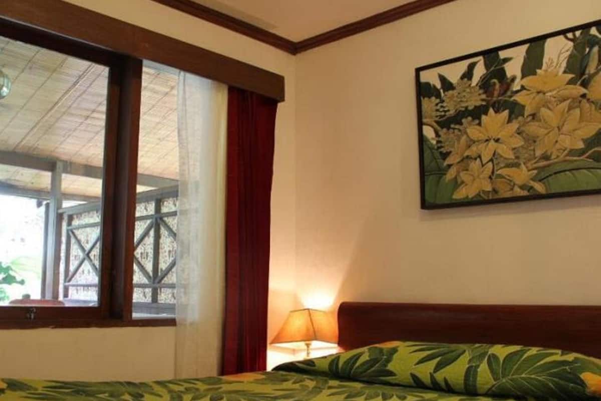 A hotel room with a bed, a window with curtains, a wall-mounted lamp, and a floral painting.