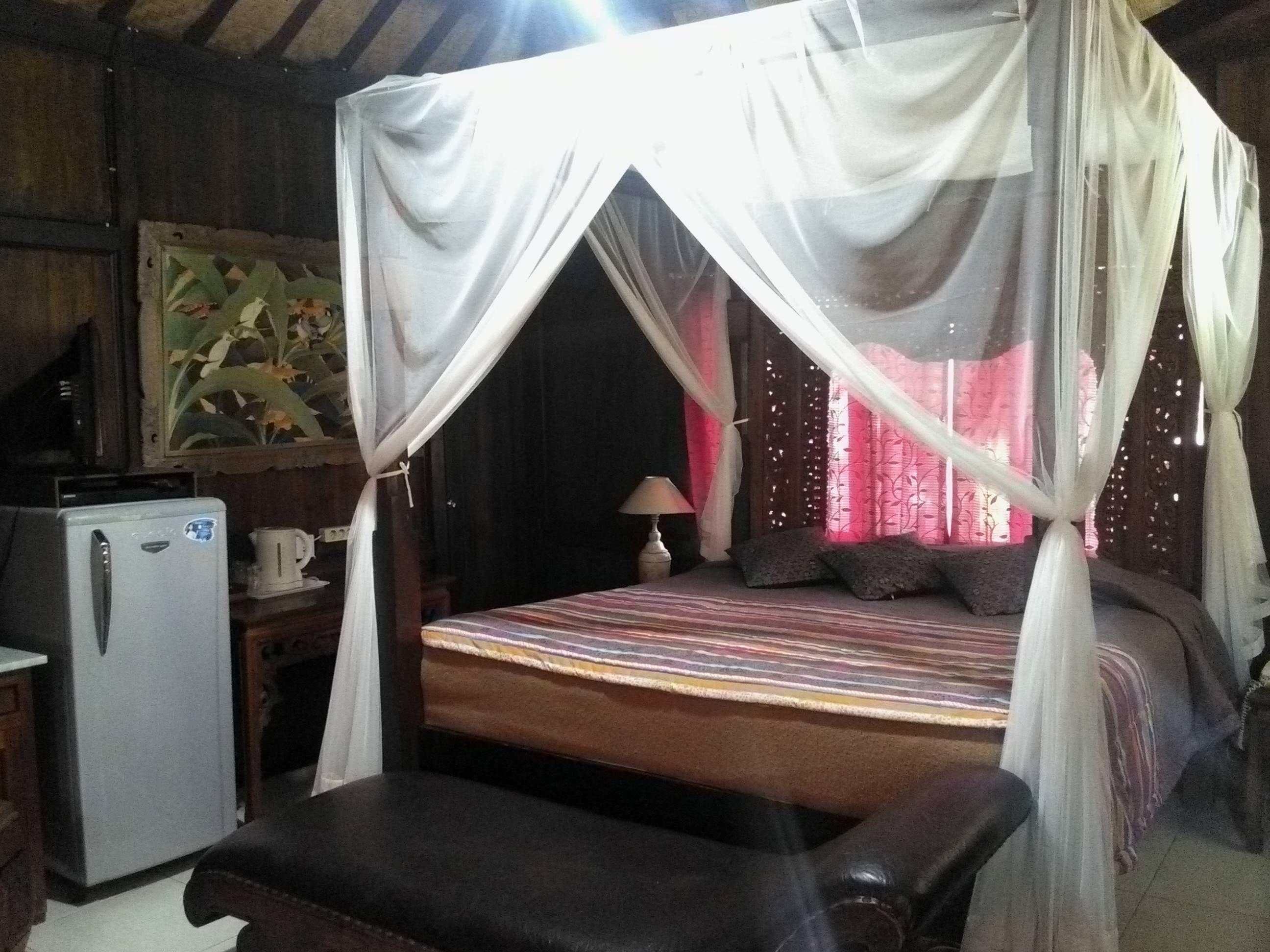 A room with a bed under a canopy, a small table with a lamp, and a refrigerator.