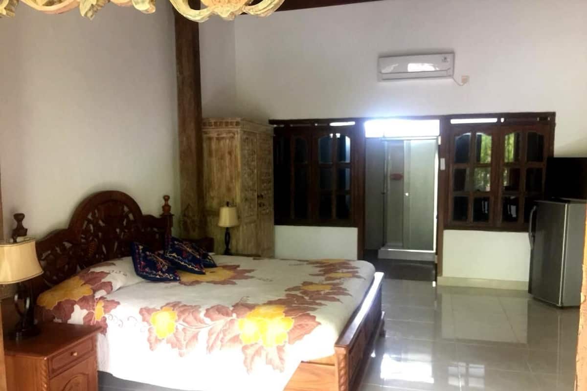 A bedroom with a bed, wooden headboard, a nightstand, a lamp, a wardrobe, and a refrigerator.