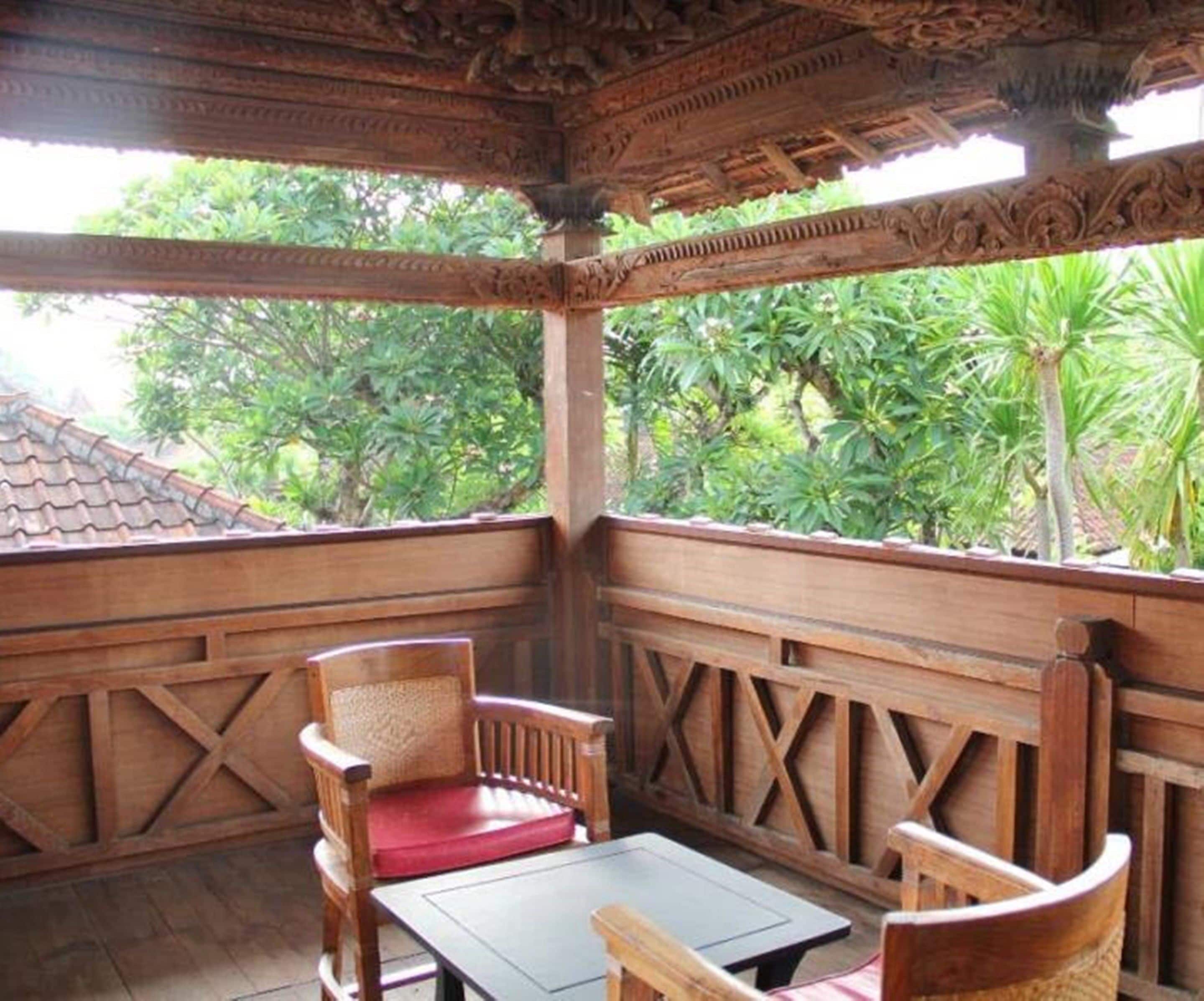 A wooden gazebo with intricate carvings, two wooden chairs with red cushions, and a table.