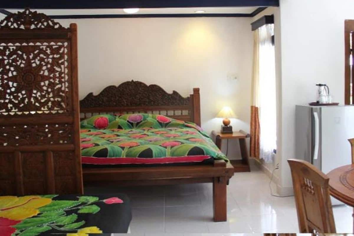 A bedroom with a wooden bed, a nightstand with a lamp, a chair, and a small table with a kettle.