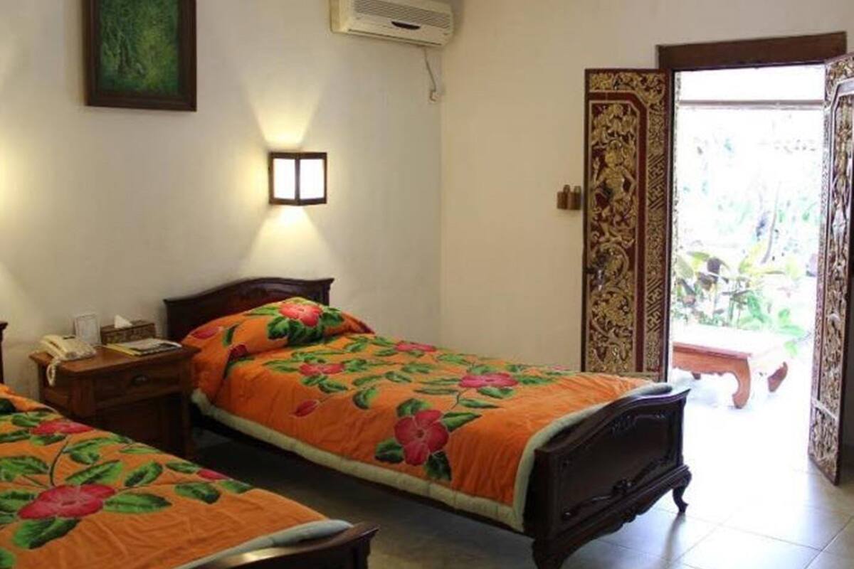 A hotel room with two single beds, a bedside table with a lamp, a wall-mounted air conditioner, and a door leading to a balcony.