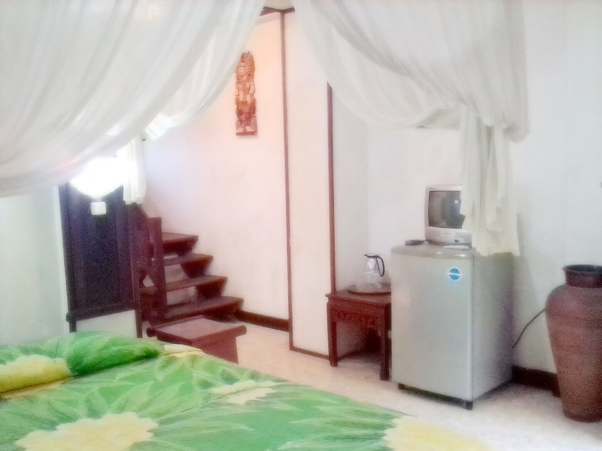 A room with a bed, a desk, a chair, a refrigerator, and a large vase.