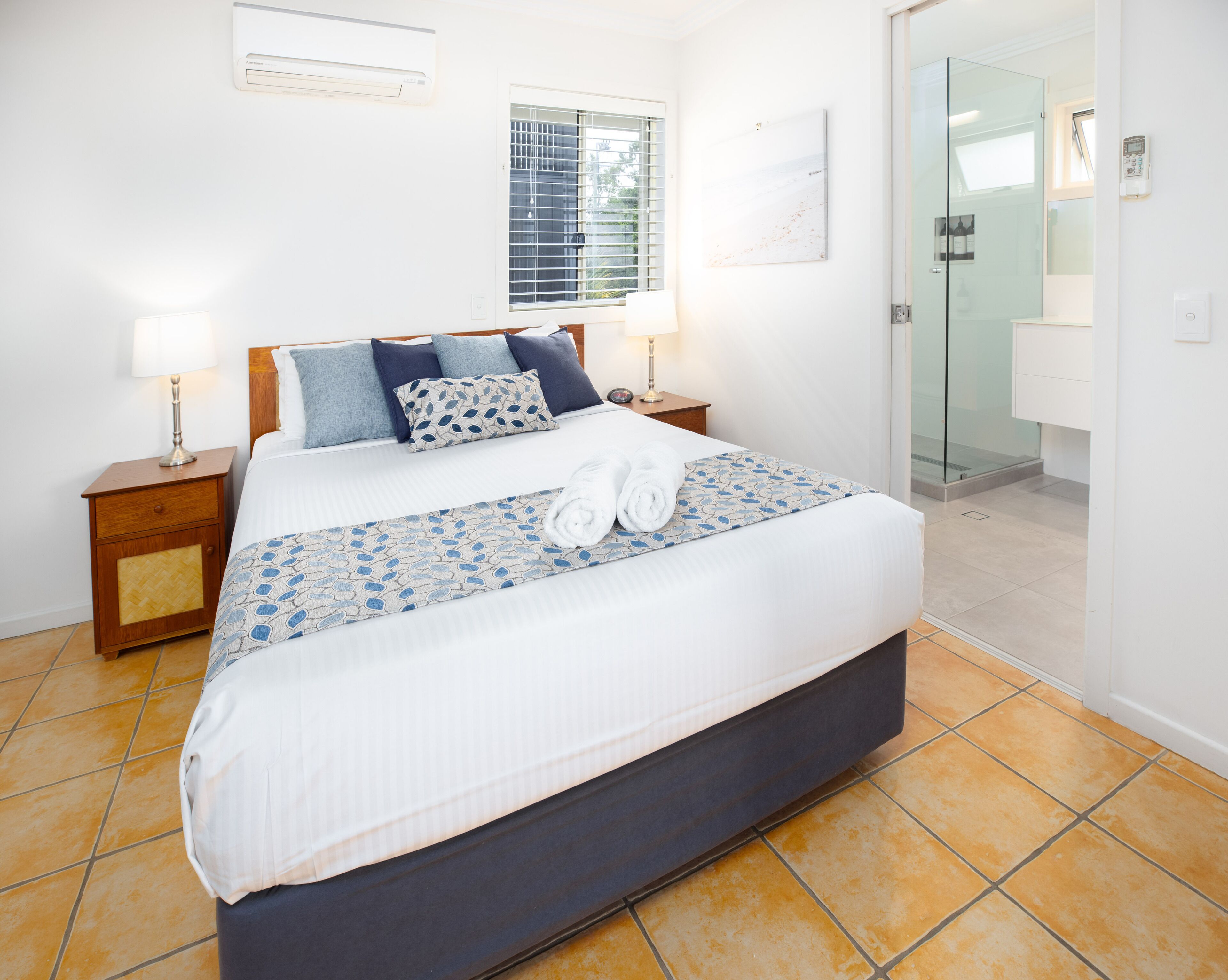 Une chambre d’hôtel comprenant un lit, deux tables de chevet, une fenêtre avec stores, un climatiseur fixé au mur, une porte en verre donnant sur une salle de bain et un sol carrelé.