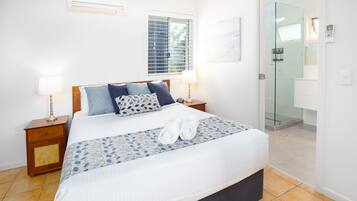 Une chambre d’hôtel comprenant un lit, deux tables de chevet, une fenêtre avec stores, un climatiseur fixé au mur, une porte en verre donnant sur une salle de bain et un sol carrelé.