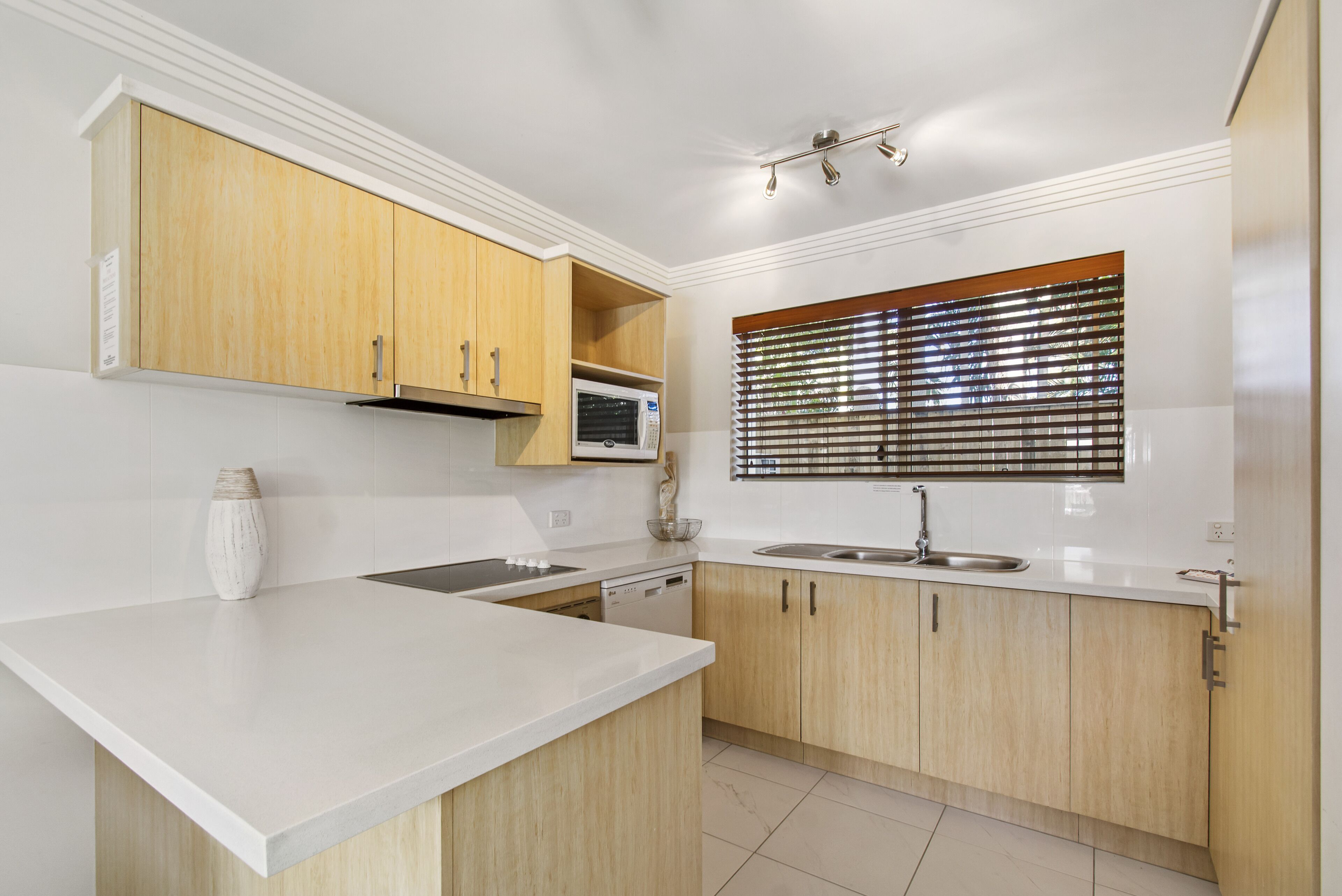 A modern kitchen with white countertops, wooden cabinets, a microwave, and a window with blinds.