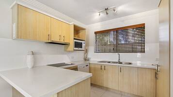 A modern kitchen with white countertops, wooden cabinets, a microwave, and a window with blinds.