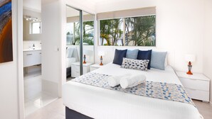 A modern bedroom with a large bed, a glass shower, and a view of palm trees.