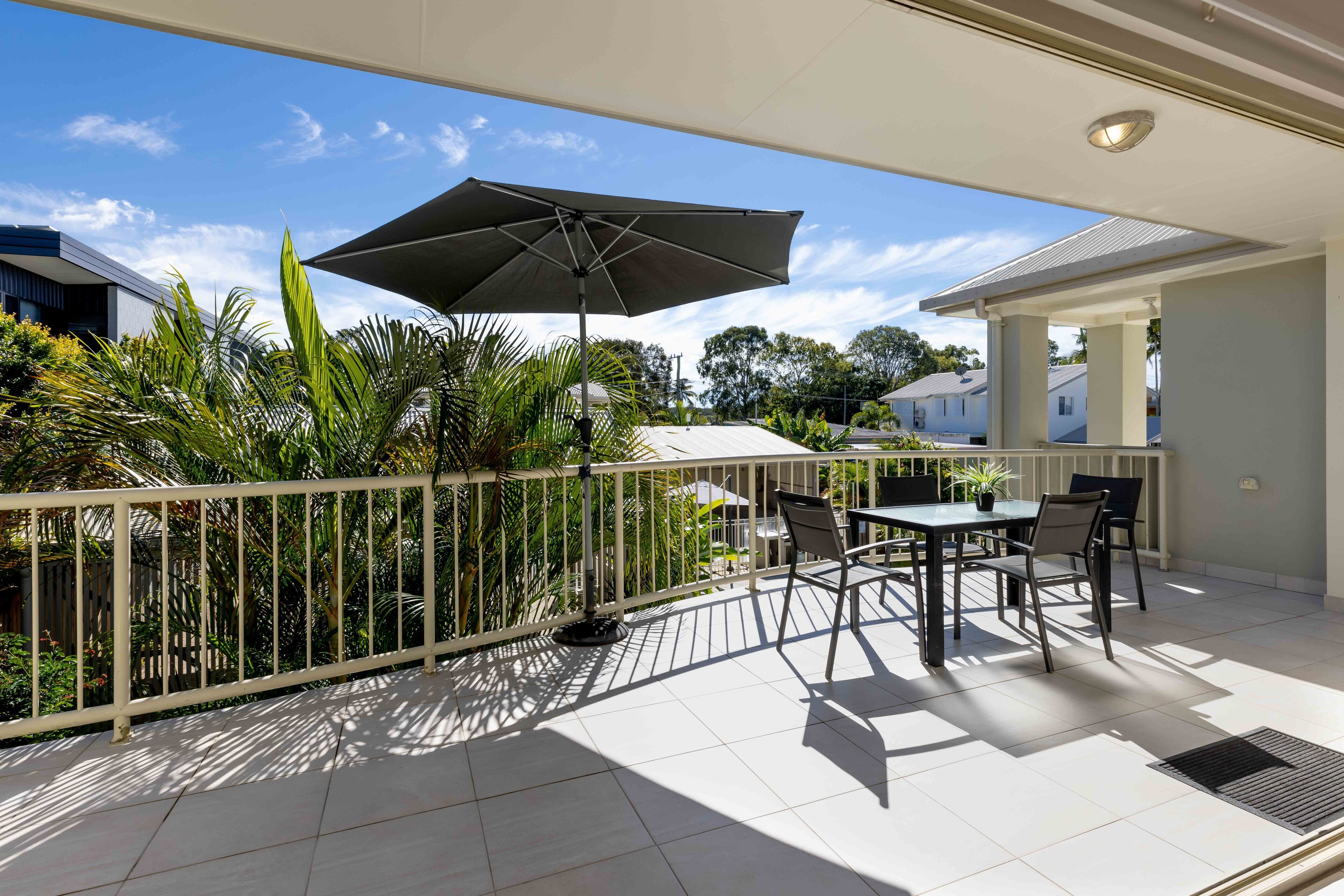 A balcony with a table and chairs, a large umbrella, and a view of surrounding houses and greenery.