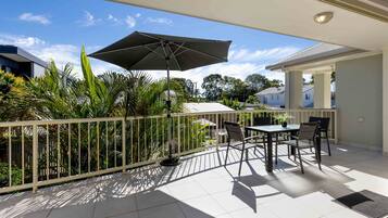A balcony with a table and chairs, a large umbrella, and a view of surrounding houses and greenery.