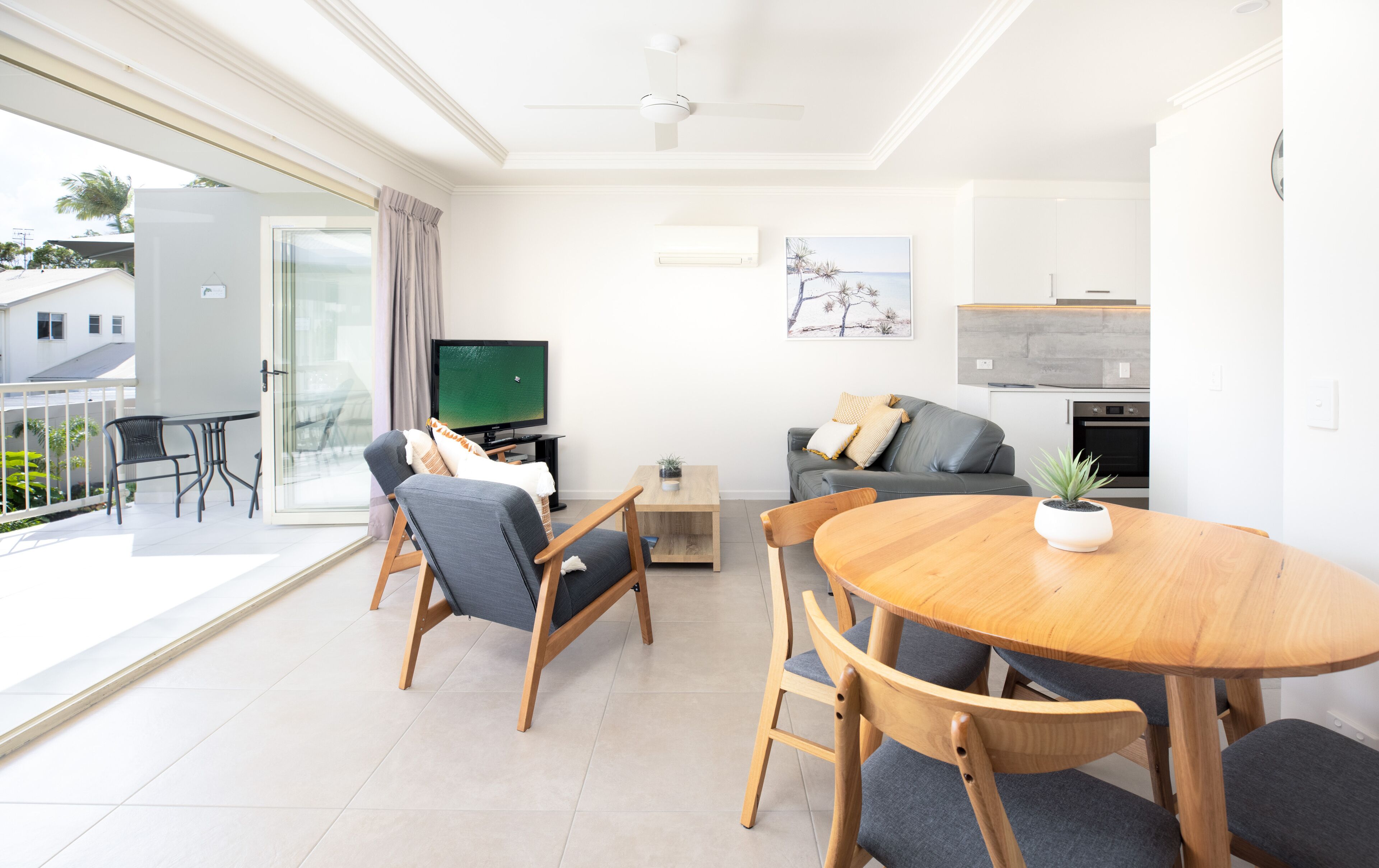Uma sala de estar moderna com uma mesa de jantar de madeira, cadeiras estofadas cinzentas, um sofá cinza com almofadas, uma televisão de tela plana, uma pequena mesa lateral de madeira com uma planta e um grande painel de vidro deslizante que dá para uma varanda.