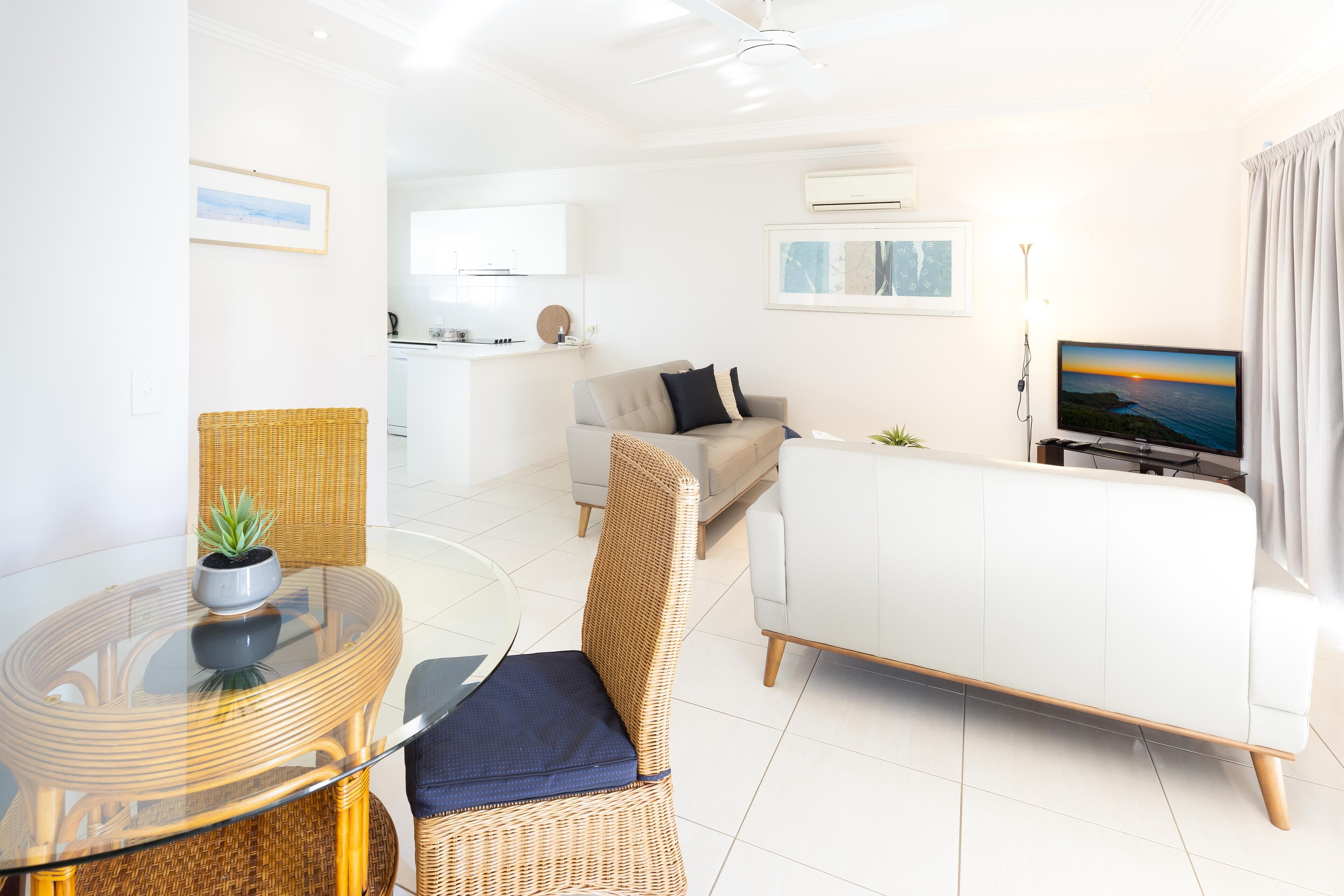 A modern living room with a glass dining table, wicker chairs, a white sofa, and a television displaying a sunset.