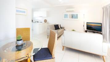 A modern living room with a glass dining table, wicker chairs, a white sofa, and a television displaying a sunset.