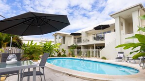A swimming pool with lounge chairs and umbrellas, surrounded by a white building with balconies.