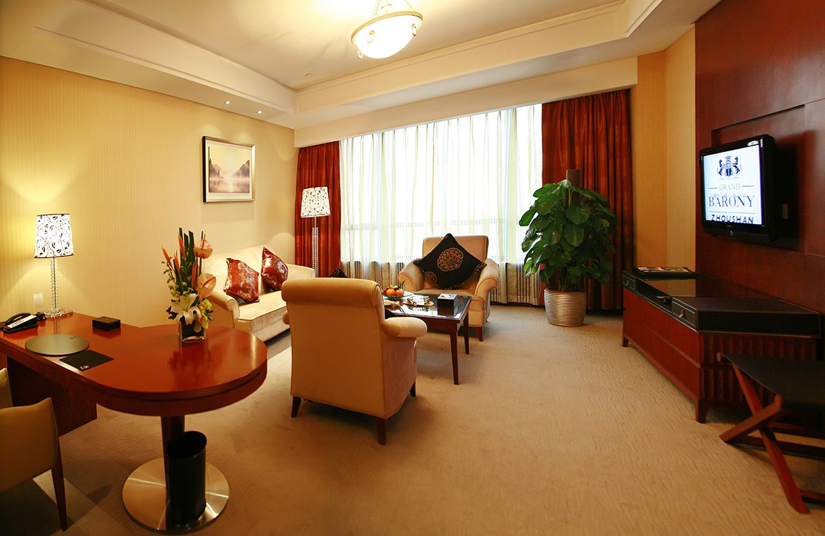 Habitación de hotel con zona de comedor, un conjunto de sofás, televisor y una planta grande.
