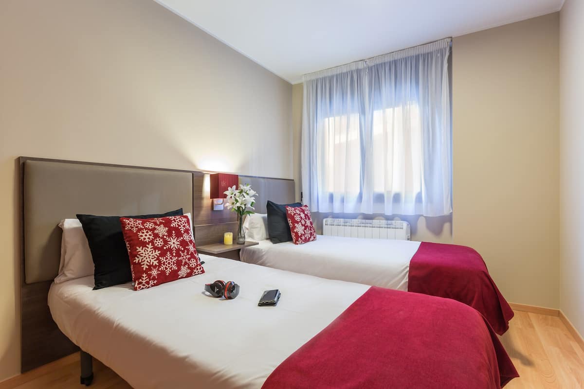 A hotel room with two beds, each with a red and white pillow, a bedside table with a lamp, and a window with sheer curtains.
