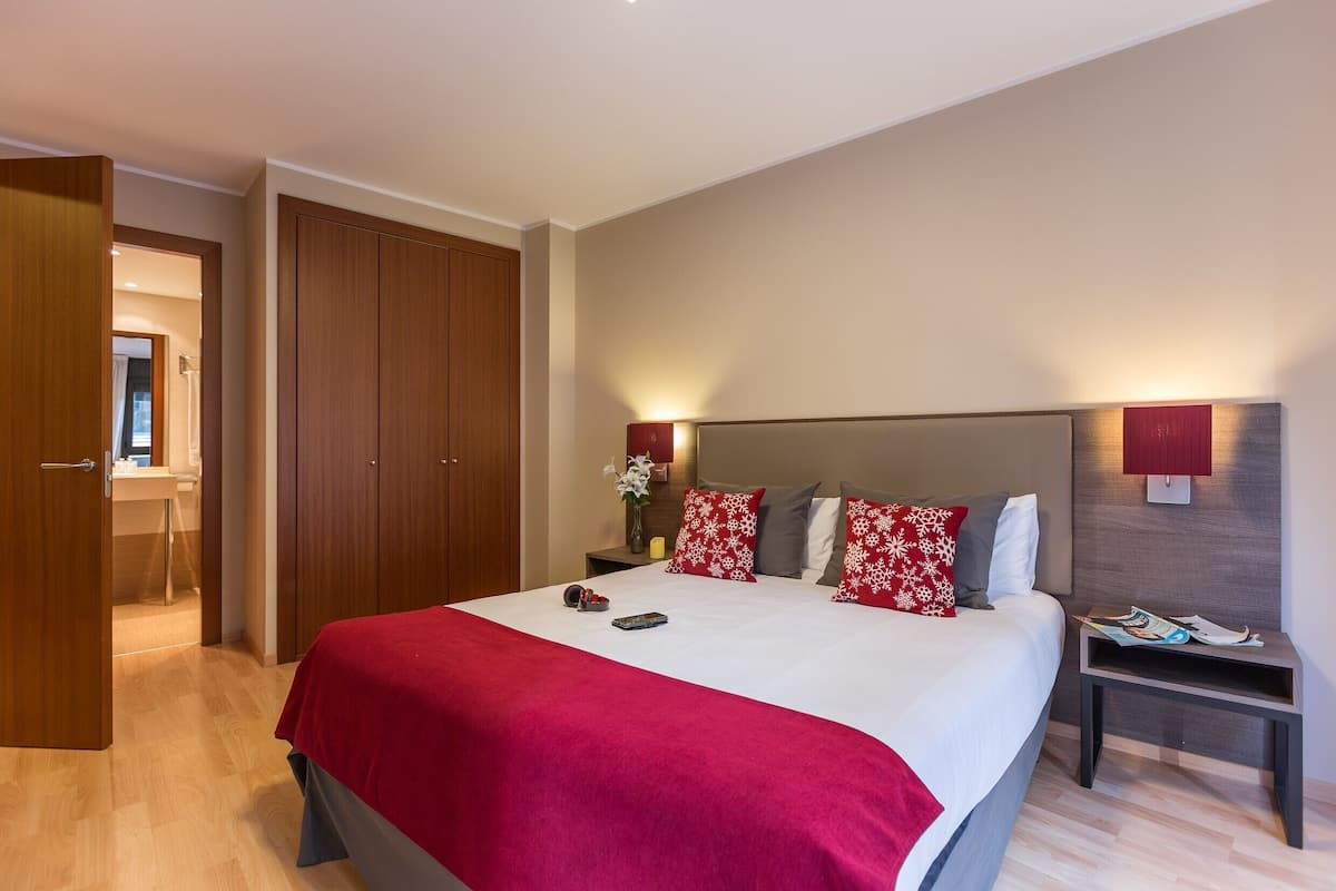 A hotel room with a large bed, wooden wardrobe, bedside table with a lamp, and a nightstand with a magazine.