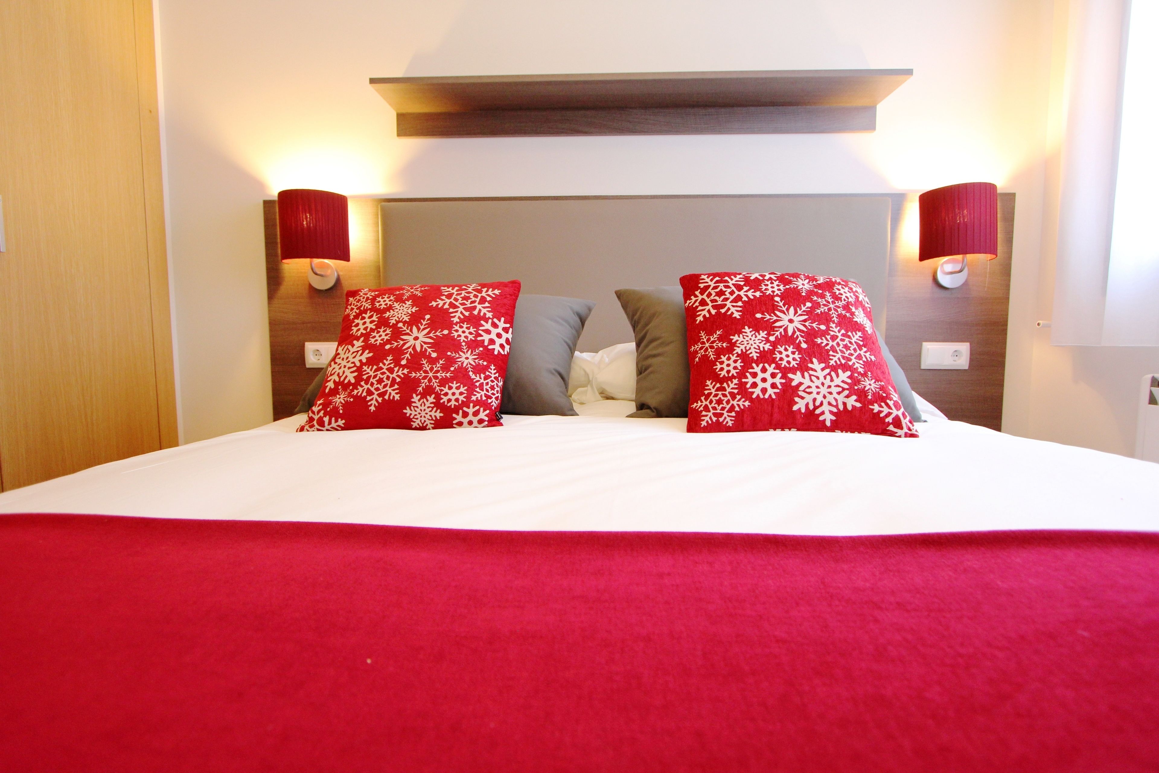 Una habitación de hotel con una cama grande, almohadas estampadas en rojo y blanco, una colcha roja y dos lámparas fijadas a la pared.