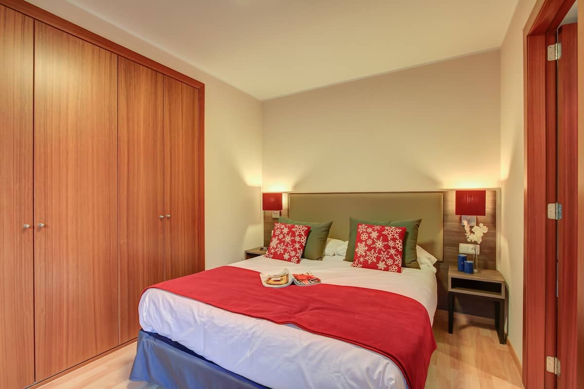 A hotel room with a large wooden wardrobe, a bed with red and white bedding, two bedside tables with lamps, and a nightstand with a vase of flowers.