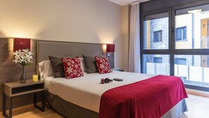 A modern hotel room with a large bed, a nightstand with a vase of flowers, and a view of the outside through a large window. - Pierre & Vacances Andorra El Tarter (El Tarter)