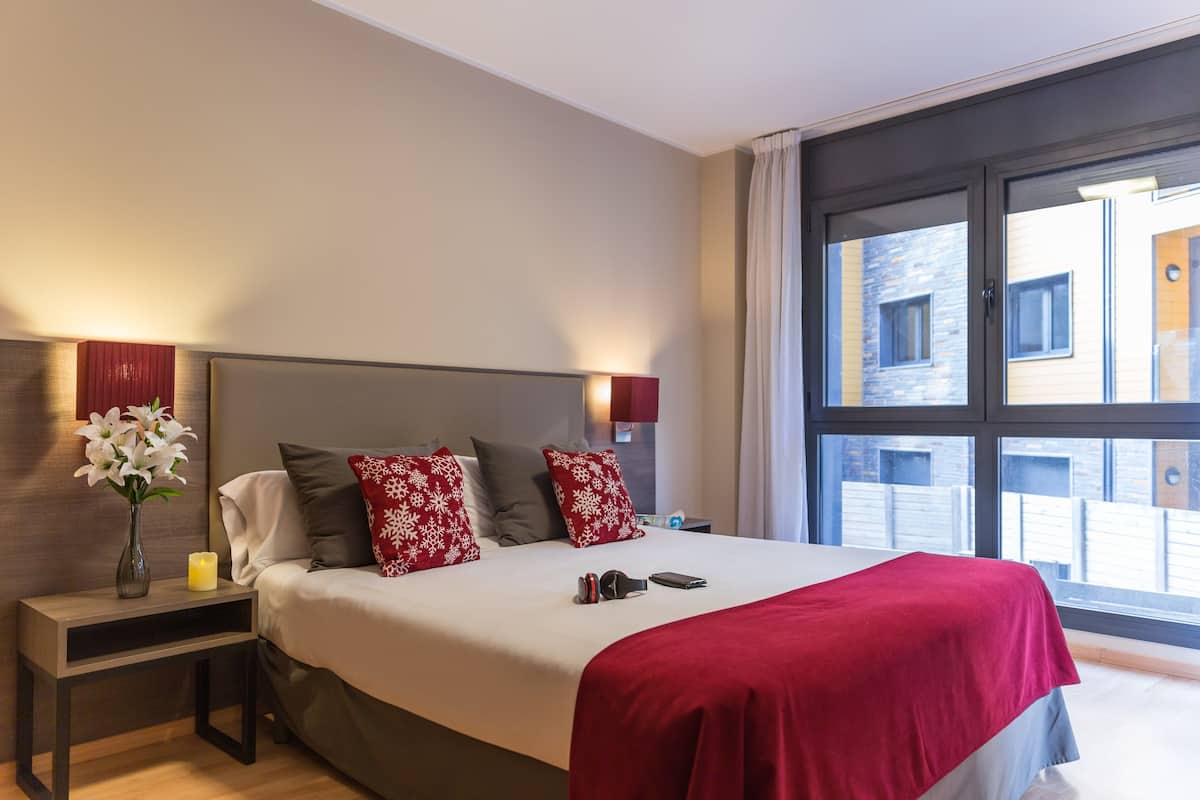 A modern hotel room with a large bed, a nightstand with a vase of flowers, and a view of the outside through a large window.