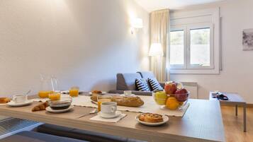 Une table dressée pour le petit-déjeuner, avec des croissants, du jus de fruits et des fruits, dans une pièce dotée d’une fenêtre et d’une lampe.