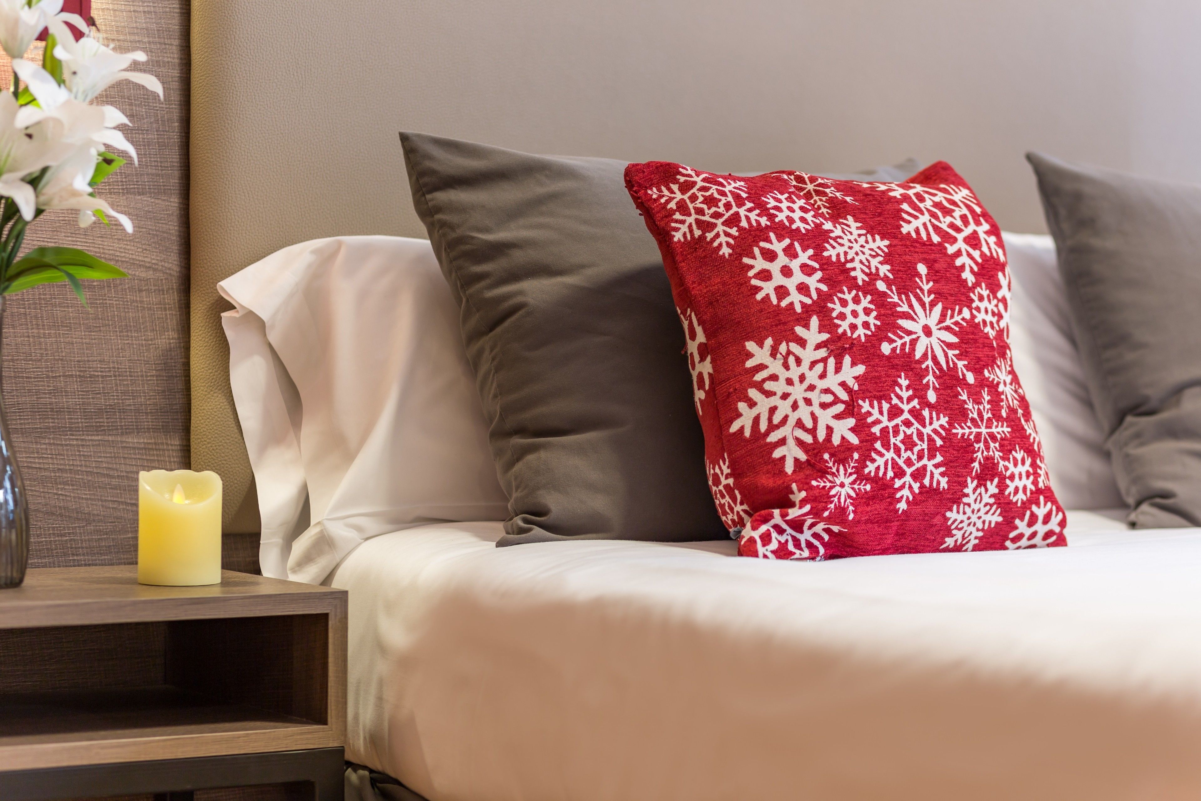 A neatly made bed with white and gray bedding, a red pillow with white snowflakes, and a lit candle on a bedside table.