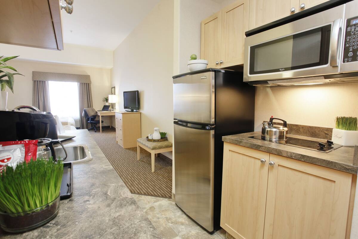 A modern kitchen with stainless steel appliances, a microwave, and a refrigerator. There is a small dining area with a table and chairs, and a window with curtains.