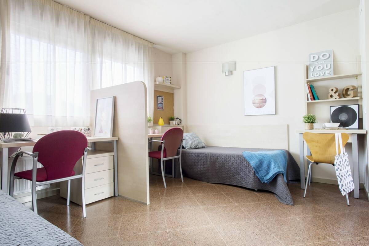A compact bedroom with a bed, desk, chair, and a small shelf with books and decorative items.