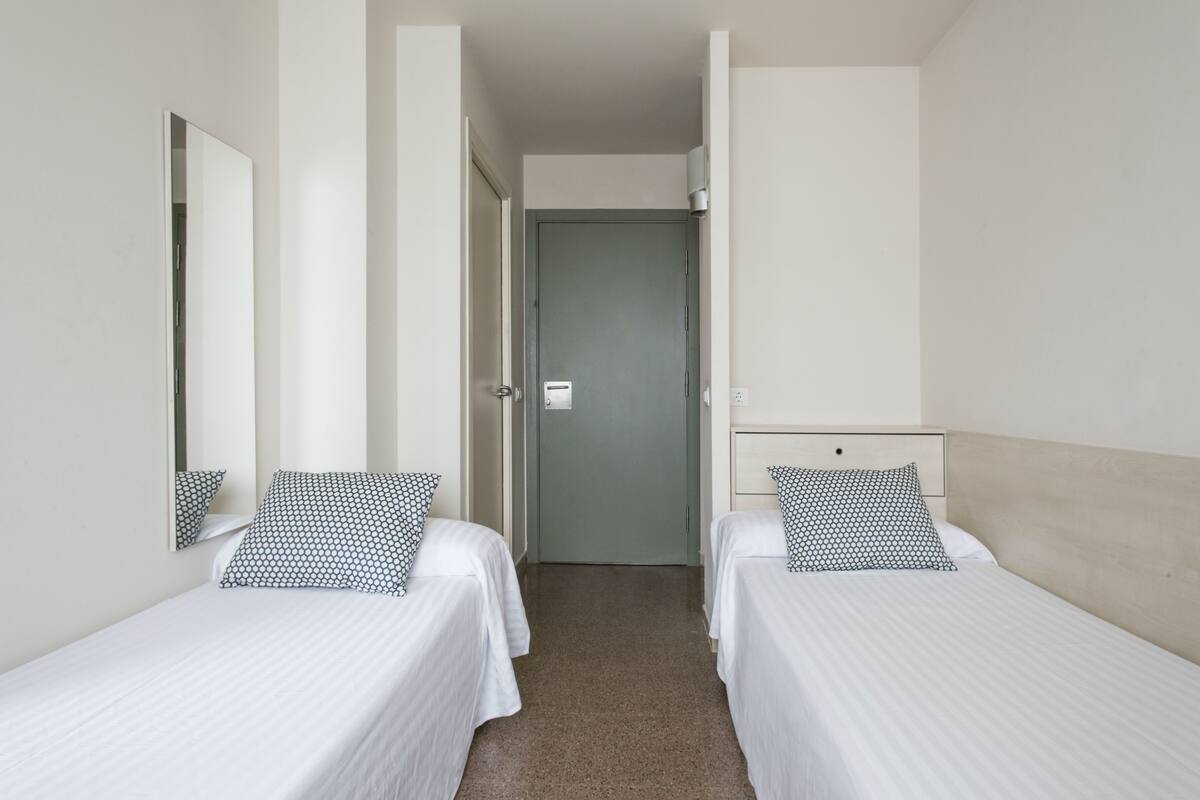 A hotel room with two single beds, a mirror, a door, and a light switch.