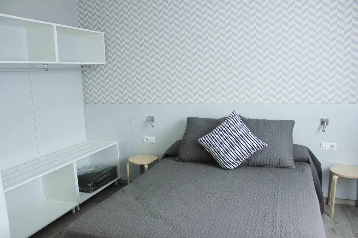 A modern bedroom with a grey bed, two bedside tables, a white wardrobe, and a patterned wallpaper.