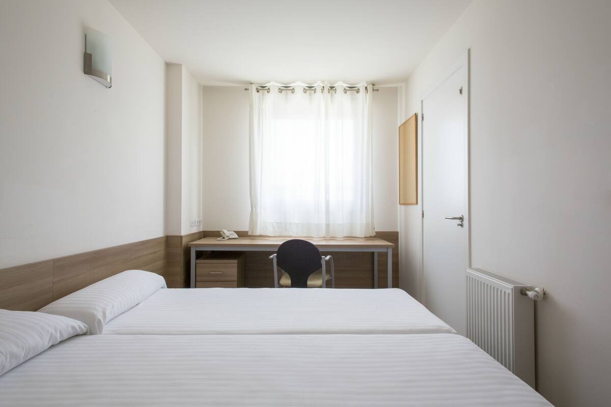 A hotel room with a bed, a desk, a chair, a window with curtains, and a door.