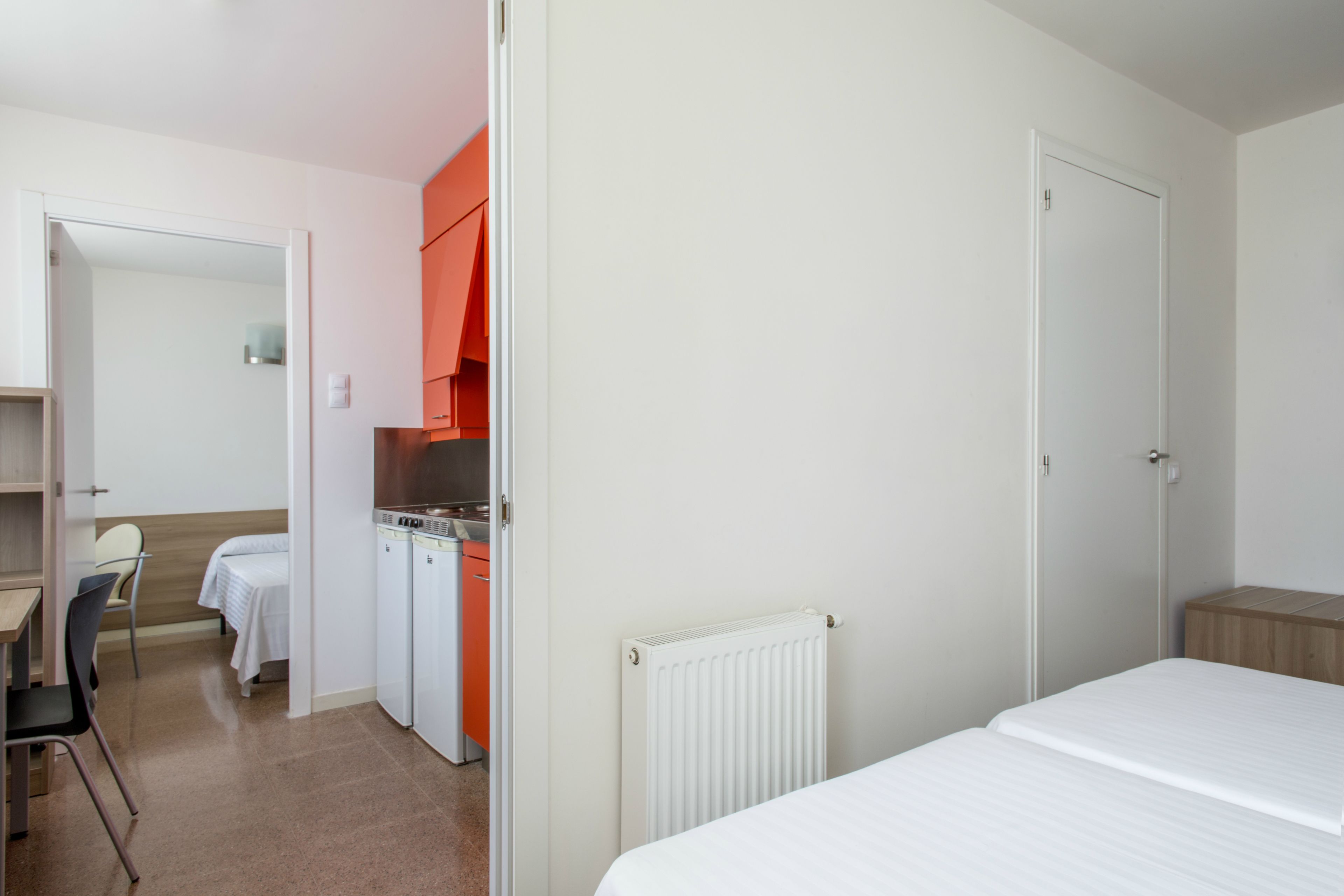 Quarto de hotel com cama, escrivaninha, cadeira, cozinha compacta e banheiro.