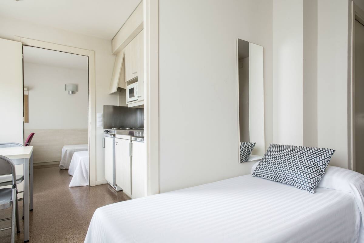 A hotel room with a bed, a desk, a chair, a microwave, and a small kitchenette.