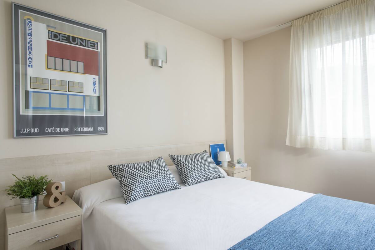 A hotel room with a large bed, two pillows, a nightstand with a lamp, a framed picture on the wall, and a window with sheer curtains.