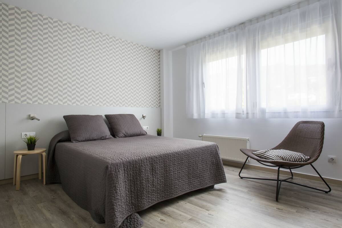 A bedroom with a bed, a chair, a small table, and a window with sheer curtains.